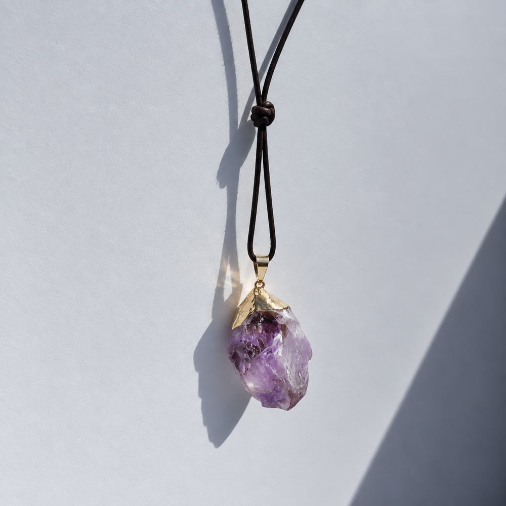 A raw purple amethyst crystal pendant necklace with a gold cap and black cord, shown against a light gray background with a strong diagonal shadow.