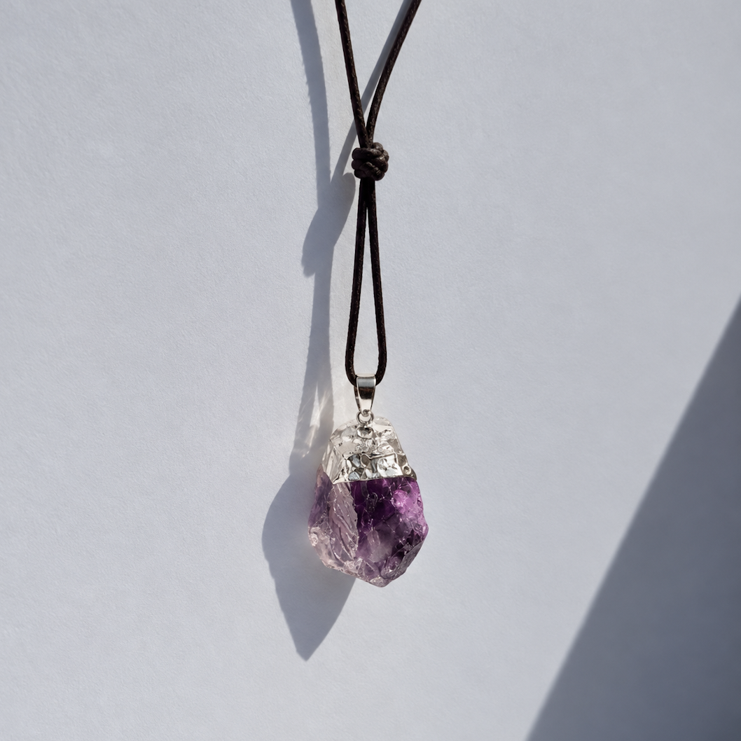 A raw purple amethyst crystal pendant necklace on a black cord, photographed against a light gray background with a strong diagonal shadow.